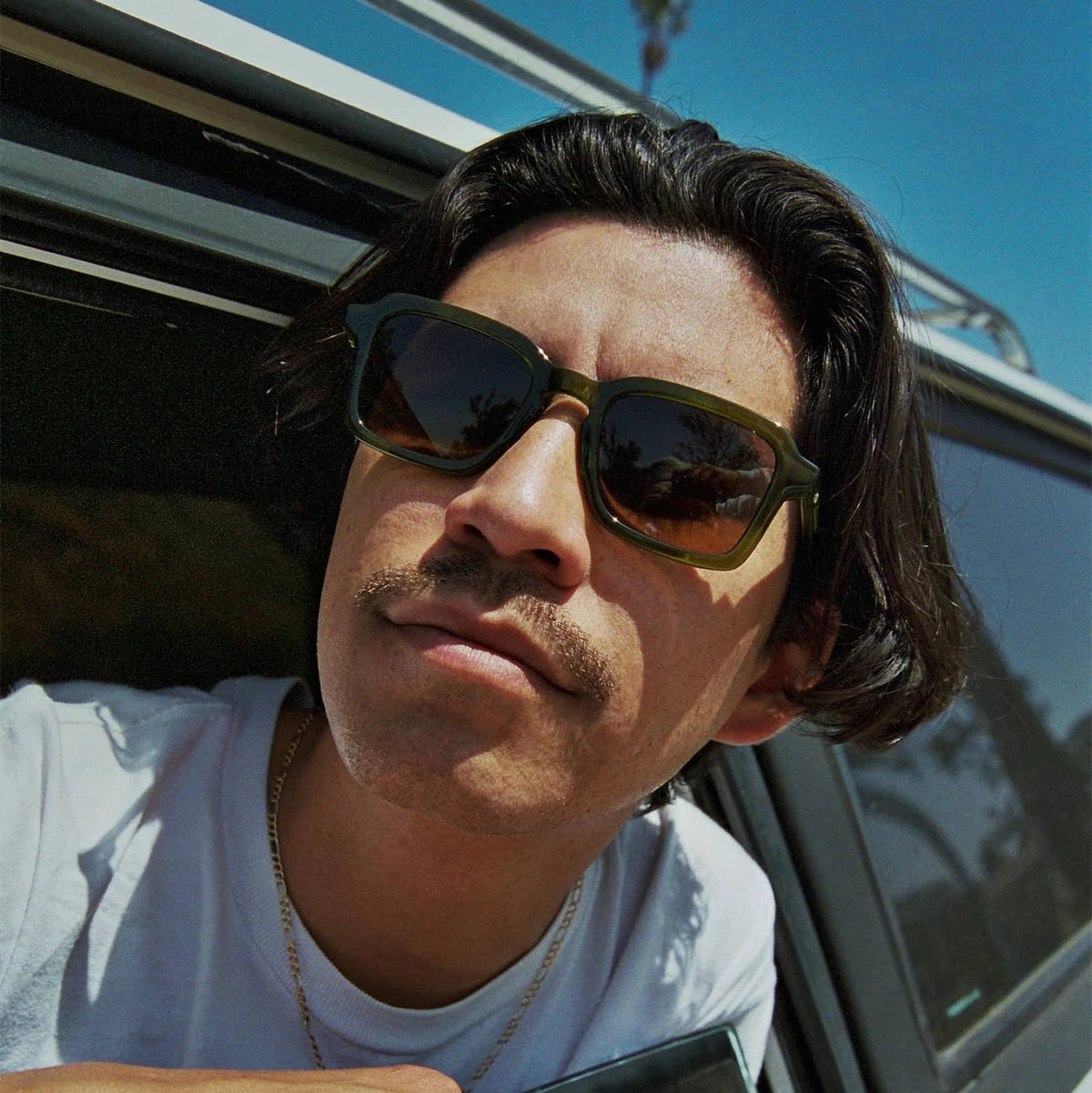 Man wearing sunglasses and a white shirt leaning out of a car window.