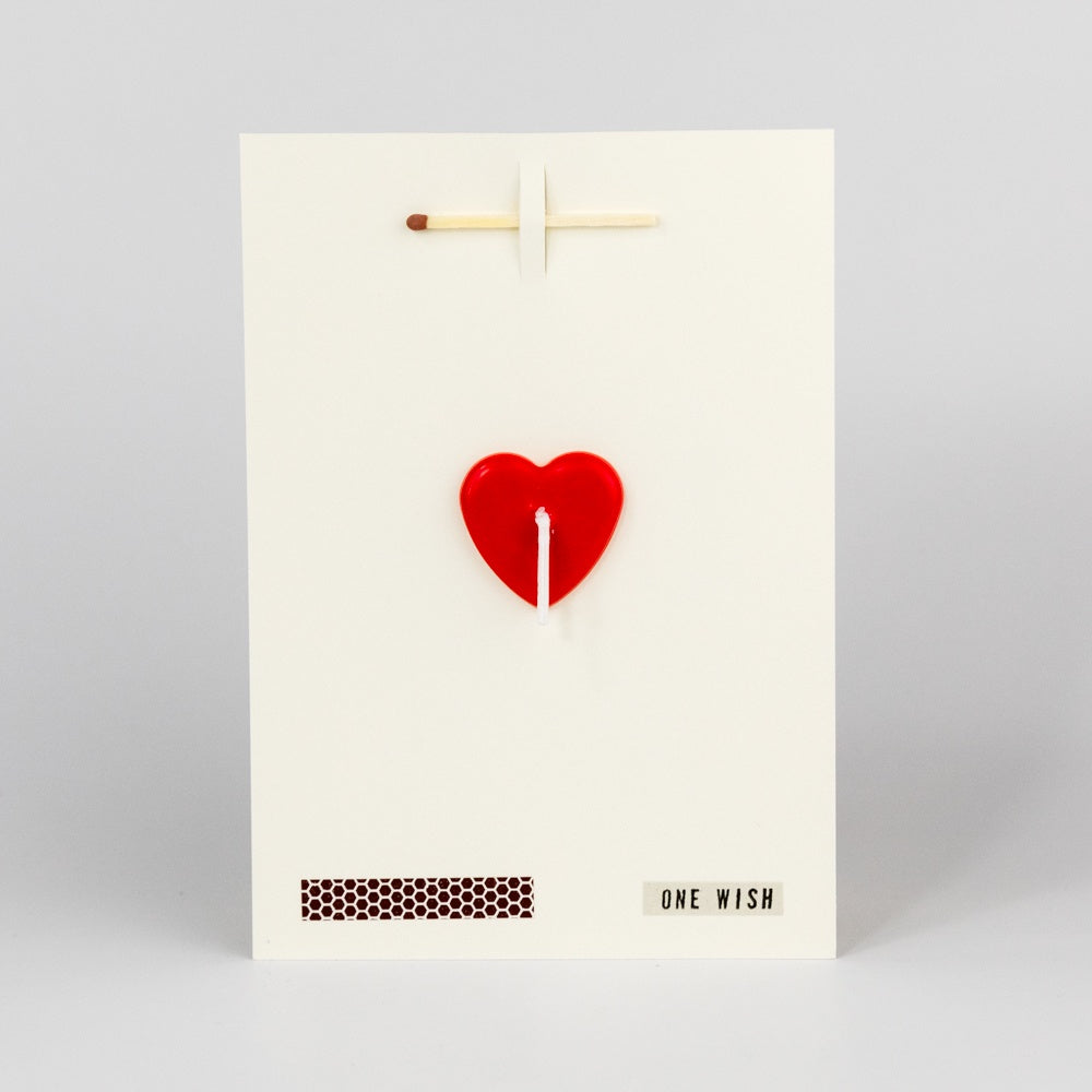 A greeting card with a red heart shape and a white background, featuring a match, a strike-pad, and a candlewick. The text 'One Wish' is printed on the card.